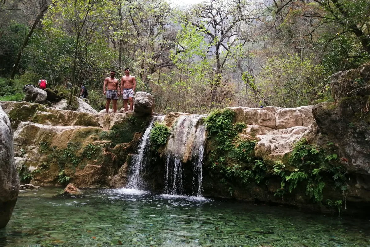 Excursionistas se asoman desde lo alto de una cascada hacia la poza turquesa de abajo