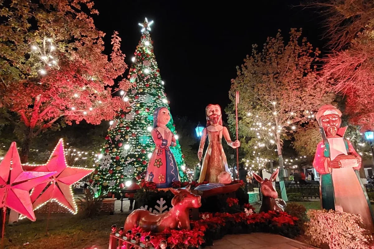 Nacimiento navideño y árbol iluminado junto a árboles con luces rojas en la plaza del pueblo